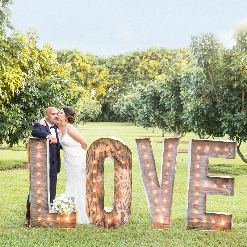 Newlyweds share a kiss in front of a glowing love sign at Longan’s Place, a dreamy South Florida venue.